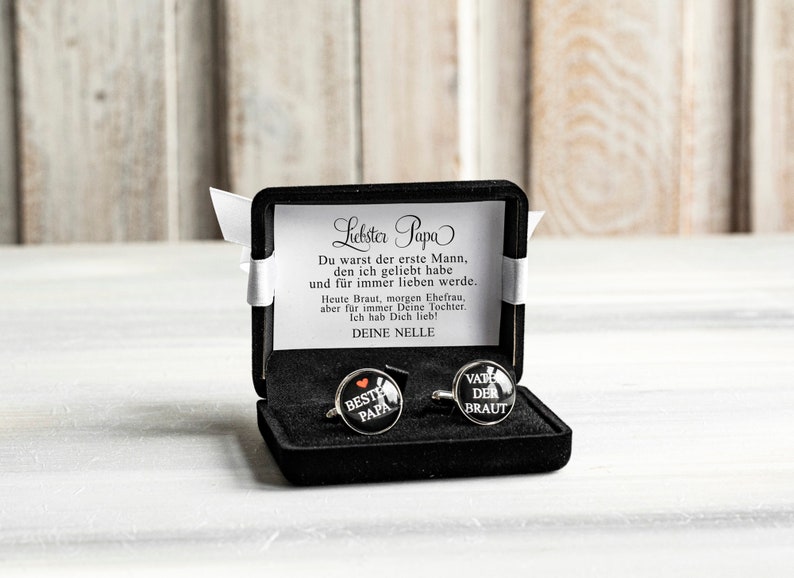 May include: A black gift box with a white satin ribbon and a card that reads "Liebster Papa Du warst der erste Mann, den ich geliebt habe und f&uuml;r immer lieben werde. Heute Braut, morgen Ehefrau, aber f&uuml;r immer Deine Tochter. Ich hab Dich lieb! DEINE NELLE". The box contains two cufflinks, one with the text "BESTE PAPA" and the other with the text "VATE DER BRAUT".