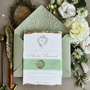 May include: A wedding invitation suite featuring a white card with gold deckled edges, a sage green envelope with a floral embossed flap, a sage green ribbon belly band, a gold wax seal with the letters AD, and a vintage gold quill pen.