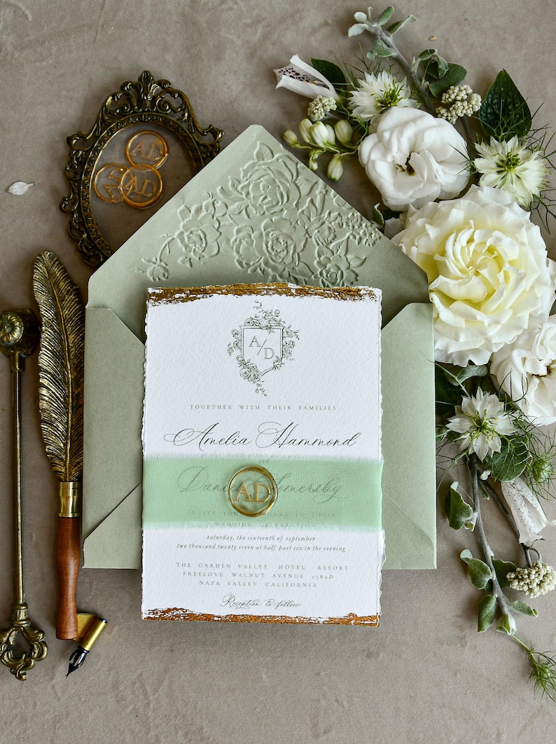 May include: A wedding invitation suite featuring a white card with gold deckled edges, a sage green envelope with a floral embossed flap, a sage green ribbon belly band, a gold wax seal with the letters AD, and a vintage gold quill pen.