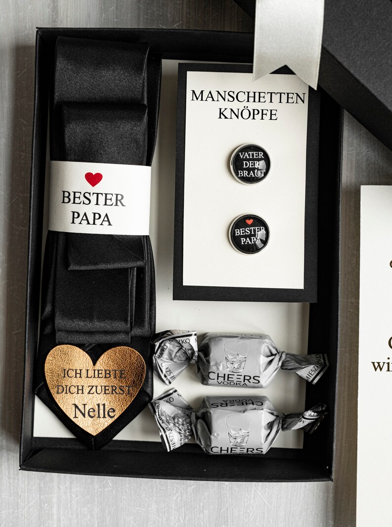 May include: A black gift box with a black tie, two cufflinks, and two chocolate candies. The cufflinks have the text "Vater der Braut" and "Bester Papa" with a red heart. The tie has a gold heart with the text "Ich liebte dich zuerst Nelle". The candies are wrapped in silver foil with the text "Cheers Vodka".