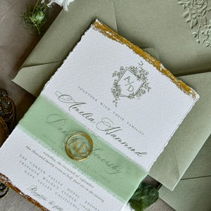 May include: A white wedding invitation with deckled edges and gold leaf accents, featuring a floral crest with the initials A/D, black calligraphy text, and a sage green band. A circular gold wax seal with the initials A/D sits on the invitation.