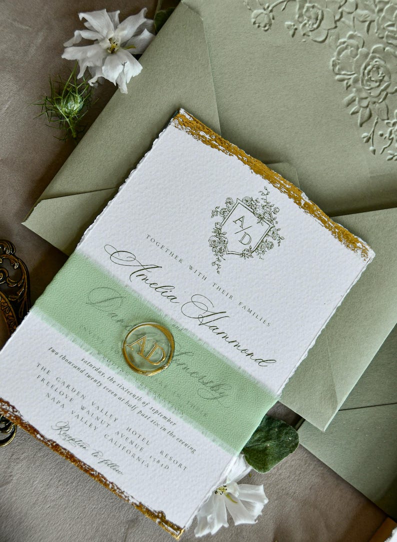 May include: A white wedding invitation with deckled edges and gold leaf accents, featuring a floral crest with the initials A/D, black calligraphy text, and a sage green band. A circular gold wax seal with the initials A/D sits on the invitation.