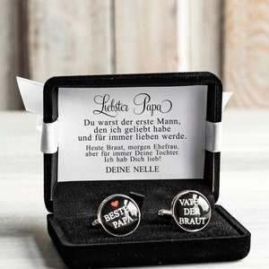 May include: A black velvet box with a white ribbon contains two cufflinks. One cufflink has a red heart and the text "BESTE PAPA" on a black background. The other cufflink has the text "VATE DER BRAUT" on a black background.
