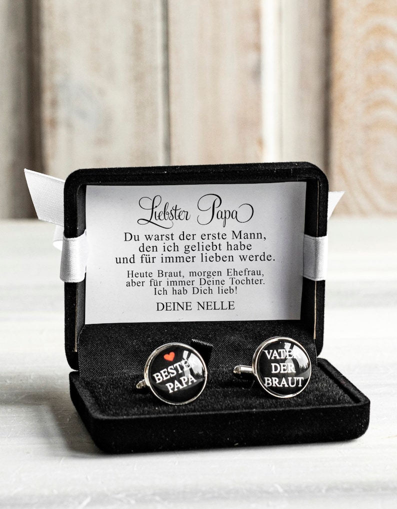 May include: A black velvet box with a white ribbon contains two cufflinks. One cufflink has a red heart and the text "BESTE PAPA" on a black background. The other cufflink has the text "VATE DER BRAUT" on a black background.