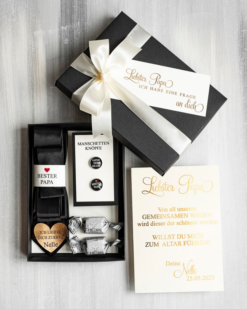 May include: A black gift box with a white satin ribbon and bow. The box contains a black bow tie, cufflinks, and a heart-shaped wooden plaque with the text "I loved you first, Nelle". The box also includes a card with the text "Dearest Dad, From all our shared journeys, this will be the most beautiful. Will you walk me down the aisle? Your Nelle, 25.05.2025".