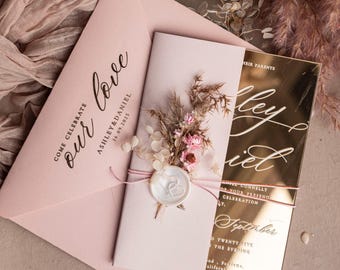 May include: A wedding invitation suite featuring a blush pink envelope with the words "our love" and "Ashley & Daniel 16.09.2025". The invitation is adorned with dried flowers and a gold mirrored card with white calligraphy.