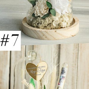May include: A glass dome with a wooden base contains a white rose, dried flowers, and greenery. A gold heart-shaped plaque with the text "F&Uuml;R IMMER UND EWIG Margo & Robert 06.06.2020" is attached to a stem inside the dome.