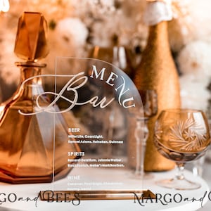 Puede incluir: Un letrero de acrílico transparente con un borde superior redondeado, que presenta el texto "MENU Bar" en una fuente cursiva. El letrero enumera varias opciones de cerveza, licores y vino. El letrero se muestra en un soporte de madera con un acabado dorado.