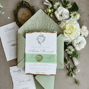 May include: A wedding invitation suite featuring a sage green envelope with a floral embossed flap, a white card with gold deckled edges, a translucent green vellum belly band, a gold wax seal with initials AD, and matching detail cards.