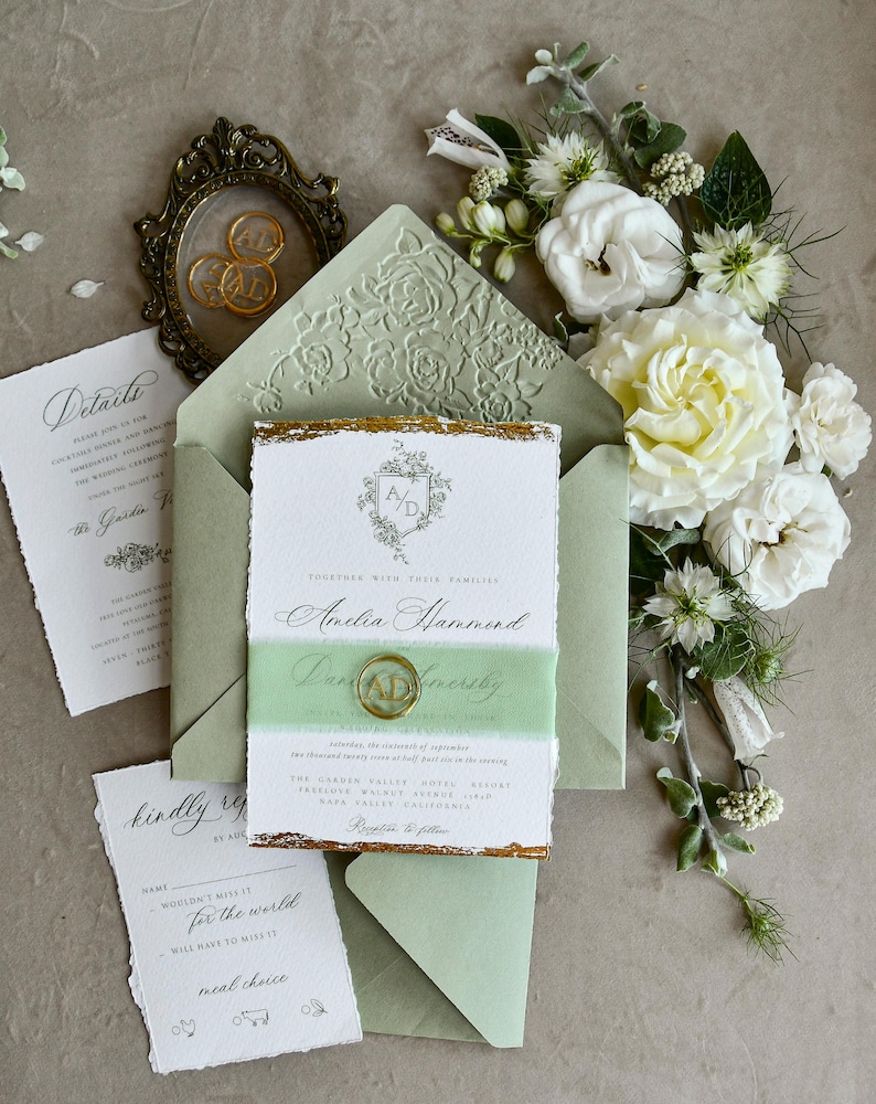 May include: A wedding invitation suite featuring a sage green envelope with a floral embossed flap, a white card with gold deckled edges, a translucent green vellum belly band, a gold wax seal with initials AD, and matching detail cards.