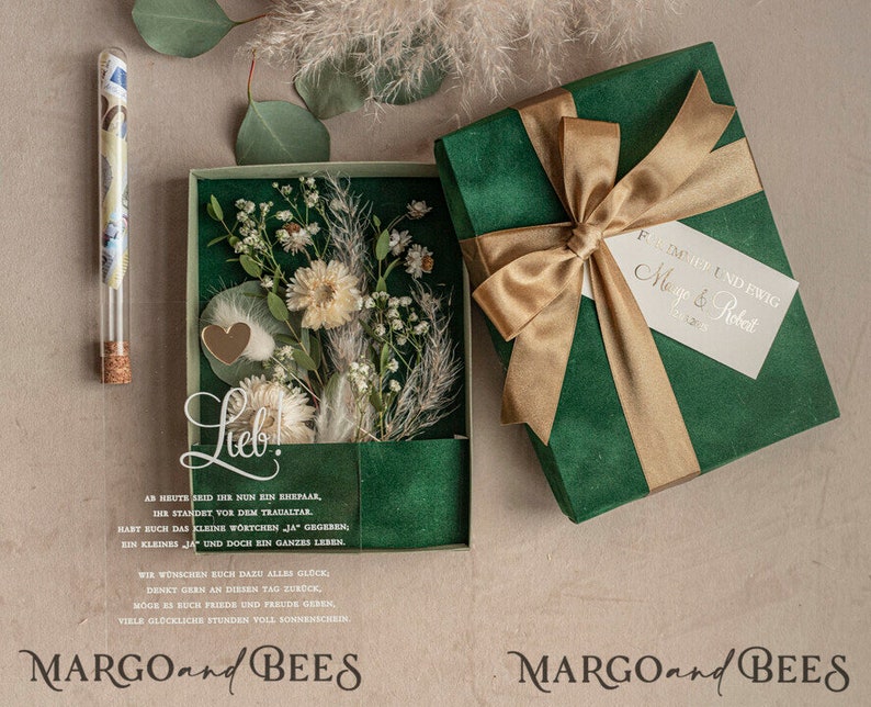 May include: A green box with a gold ribbon and a white tag that says "Forever and Ever Margo & Robert". The box is open and inside is a clear box with dried flowers and a small glass vial. The text "Lieb!" is written on the clear box. The text "MARGO and BEES" is written on the bottom of the image.