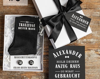 May include: A white gift box with a black ribbon and a black card that says "Alexander, Hole deinen Anzug raus - Du wirst gebraucht, Willst do mein Trauzeuge sein?" The box contains a pair of black socks with the text "Trauzeuge Bester Mann" and a pair of cufflinks with the text "Bester Mann" and "Trauzeuge".