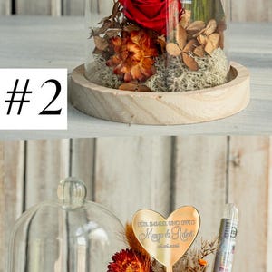 May include: A glass dome with a wooden base contains a preserved red rose and dried flowers. A gold heart-shaped plaque with the text "F&Uuml;R IMMER UND EWIG Margo & Robert 06.06.2016" is attached to a stick in the center of the dome.