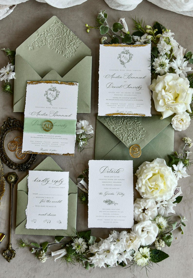 May include: A wedding stationery set featuring white paper cards with deckled edges, gold foil accents, and sage green envelopes with embossed floral patterns. The cards include text for Amelia Hammond and Daniel Somersby, a details card, and a reply card.