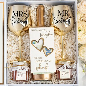 May include: A white gift box filled with a wedding gift set. The set includes two gold-rimmed wine glasses with the text "MRS Schmidt" and "MR Schmidt", a bottle of wine, a card with the text "Mit den besten W&uuml;nschen f&uuml;r eure gemeinsame Zukunft", and two small jars of jam with the text "Liebe ist s&uuml;&szlig;".