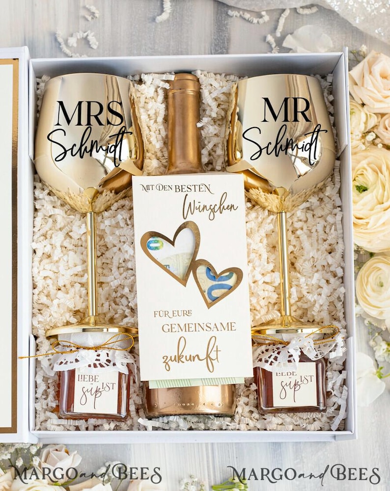 May include: A white gift box filled with a wedding gift set. The set includes two gold-rimmed wine glasses with the text "MRS Schmidt" and "MR Schmidt", a bottle of wine, a card with the text "Mit den besten W&uuml;nschen f&uuml;r eure gemeinsame Zukunft", and two small jars of jam with the text "Liebe ist s&uuml;&szlig;".