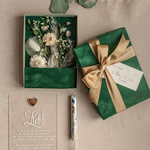 May include: A green velvet gift box with a gold ribbon and a white card that says "For immer und ewig" and "Margo & Fred" with the date "12.03.2025".  The box is open and there is a clear acrylic card with the text "Lieb!" and a small gold heart on top.  The card has a message about a wedding and the text "MARGOandBEES".  There is a small glass tube with a cork stopper and a rolled up piece of paper inside.