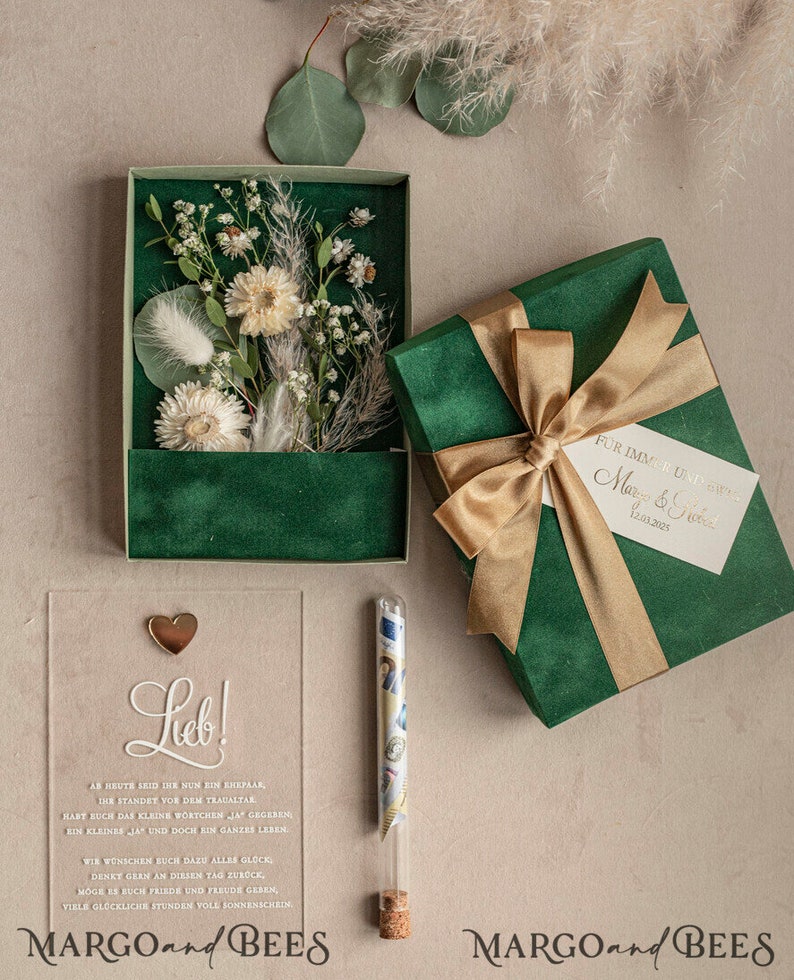 May include: A green velvet gift box with a gold ribbon and a white card that says "For immer und ewig" and "Margo & Fred" with the date "12.03.2025".  The box is open and there is a clear acrylic card with the text "Lieb!" and a small gold heart on top.  The card has a message about a wedding and the text "MARGOandBEES".  There is a small glass tube with a cork stopper and a rolled up piece of paper inside.
