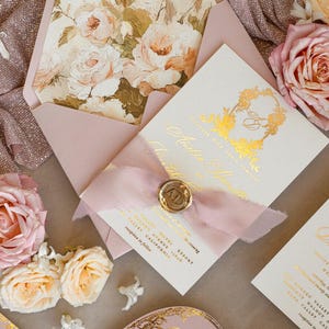May include: A wedding invitation suite with a pink envelope, a white card with gold foil lettering, and a pink ribbon with a wax seal. The invitation features a floral design with a gold monogram.