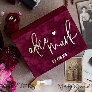 Può includere: Una scatola di velluto bordeaux con scritte dorate che recitano "Alice & Mark" e la data "13 08 23". La scatola è decorata con un piccolo cuore e si trova accanto a un portagioie in velluto e foto d'epoca.