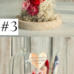 May include: A glass dome with a wooden base containing a preserved flower arrangement. The arrangement includes red, pink, and white flowers, as well as green foliage. A gold heart-shaped plaque with the text "F&uuml;r immer und ewig Margo & Robert 05.06.2026" is attached to the arrangement.