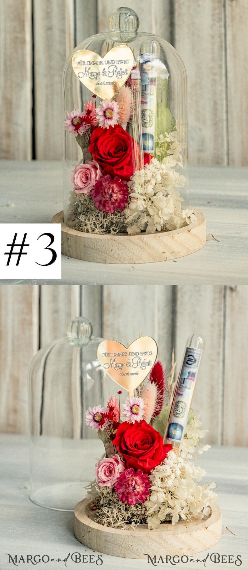 May include: A glass dome with a wooden base containing a preserved flower arrangement. The arrangement includes red, pink, and white flowers, as well as green foliage. A gold heart-shaped plaque with the text "F&uuml;r immer und ewig Margo & Robert 05.06.2026" is attached to the arrangement.