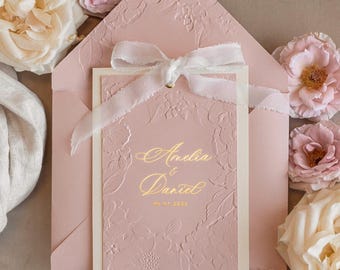 Puede incluir: Una invitación de boda rosa con una cinta blanca y letras doradas. La invitación dice "Amelia & Daniel" y "05.07.2023".