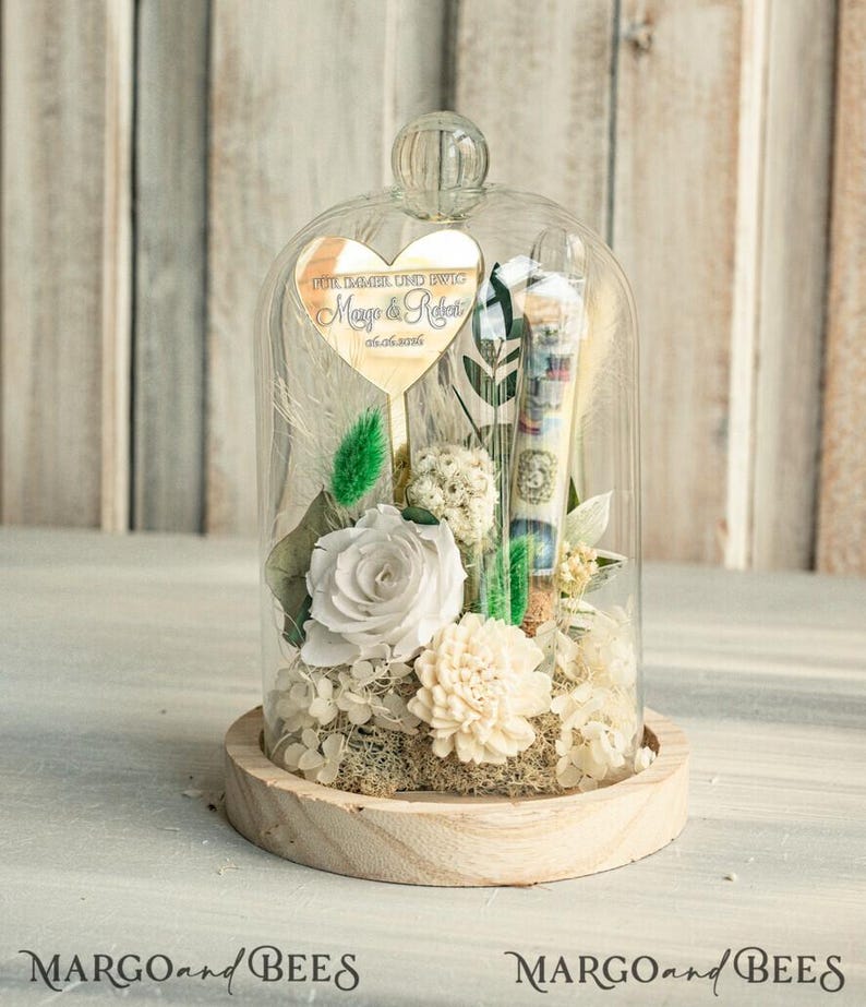 May include: A glass dome with a wooden base contains preserved white flowers and greenery. A heart-shaped mirror with the text "F&uuml;r immer und ewig Maya & Robert 05.05.2018" is attached to the inside of the dome.