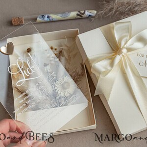 May include: A clear acrylic wedding invitation with a gold heart and the word "Lieb!" written in cursive. The invitation is decorated with dried flowers and is placed in a white box with a cream-coloured ribbon and a tag that reads "F&uuml;r immer und ewig Margo & Robert 12.03.23".