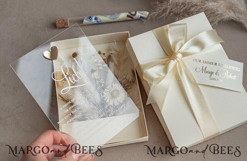 May include: A clear acrylic wedding invitation with a gold heart and the word "Lieb!" written in cursive. The invitation is decorated with dried flowers and is placed in a white box with a cream-coloured ribbon and a tag that reads "F&uuml;r immer und ewig Margo & Robert 12.03.23".