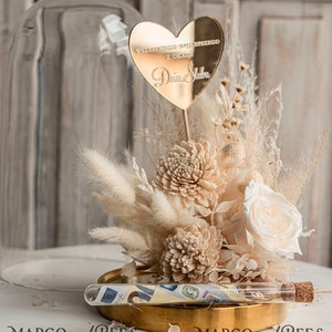 Op de afbeelding: Een glazen stolp met een gouden basis en een boeket gedroogde bloemen erin. De bloemen zijn wit en beige en omvatten pampasgras, rozen en andere gedroogde bloemen. Een gouden hartvormige plaquette met de tekst "Wszystkiego najlepszego z okazji Dnia Slubu" is bovenop het boeket geplaatst. Een glazen buis met een kurkstop bevat opgerold geld.