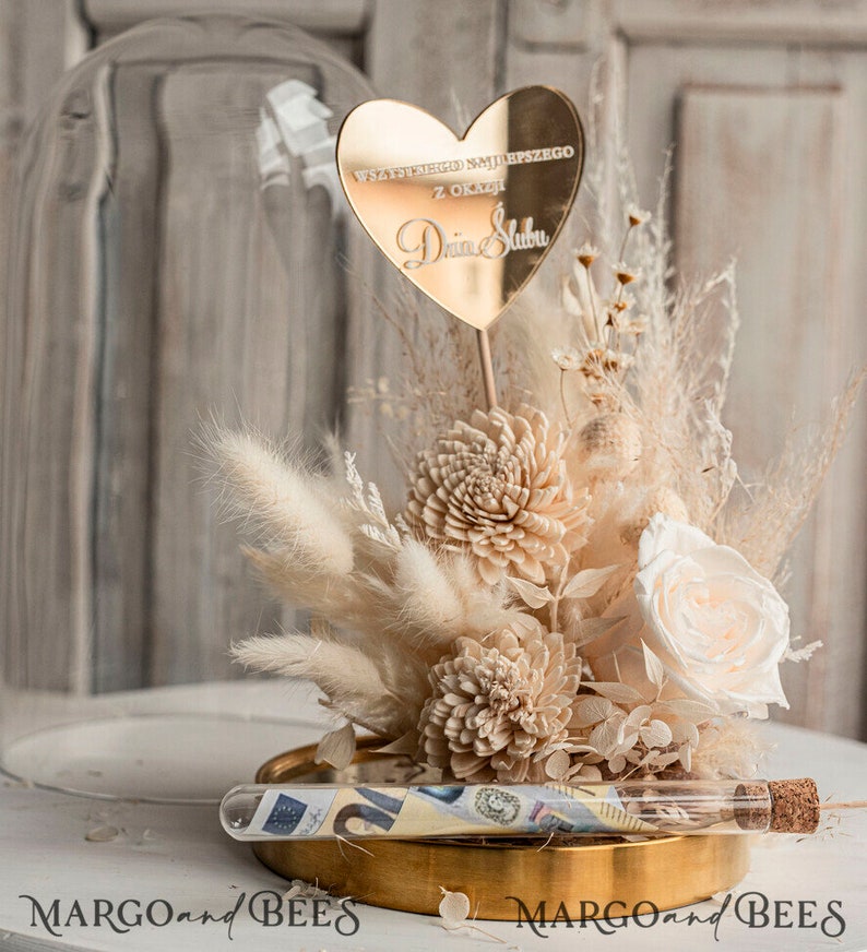 Op de afbeelding: Een glazen stolp met een gouden basis en een boeket gedroogde bloemen erin. De bloemen zijn wit en beige en omvatten pampasgras, rozen en andere gedroogde bloemen. Een gouden hartvormige plaquette met de tekst "Wszystkiego najlepszego z okazji Dnia Slubu" is bovenop het boeket geplaatst. Een glazen buis met een kurkstop bevat opgerold geld.