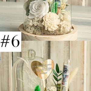 May include: A glass dome with a wooden base contains a white rose, white carnations, and green foliage. A gold heart-shaped plaque with the text "F&Uuml;R IMMER UND EWIG Mango & Relet 06.06.2016" is attached to a stick in the center of the dome. A clear glass tube filled with money is also inside the dome.