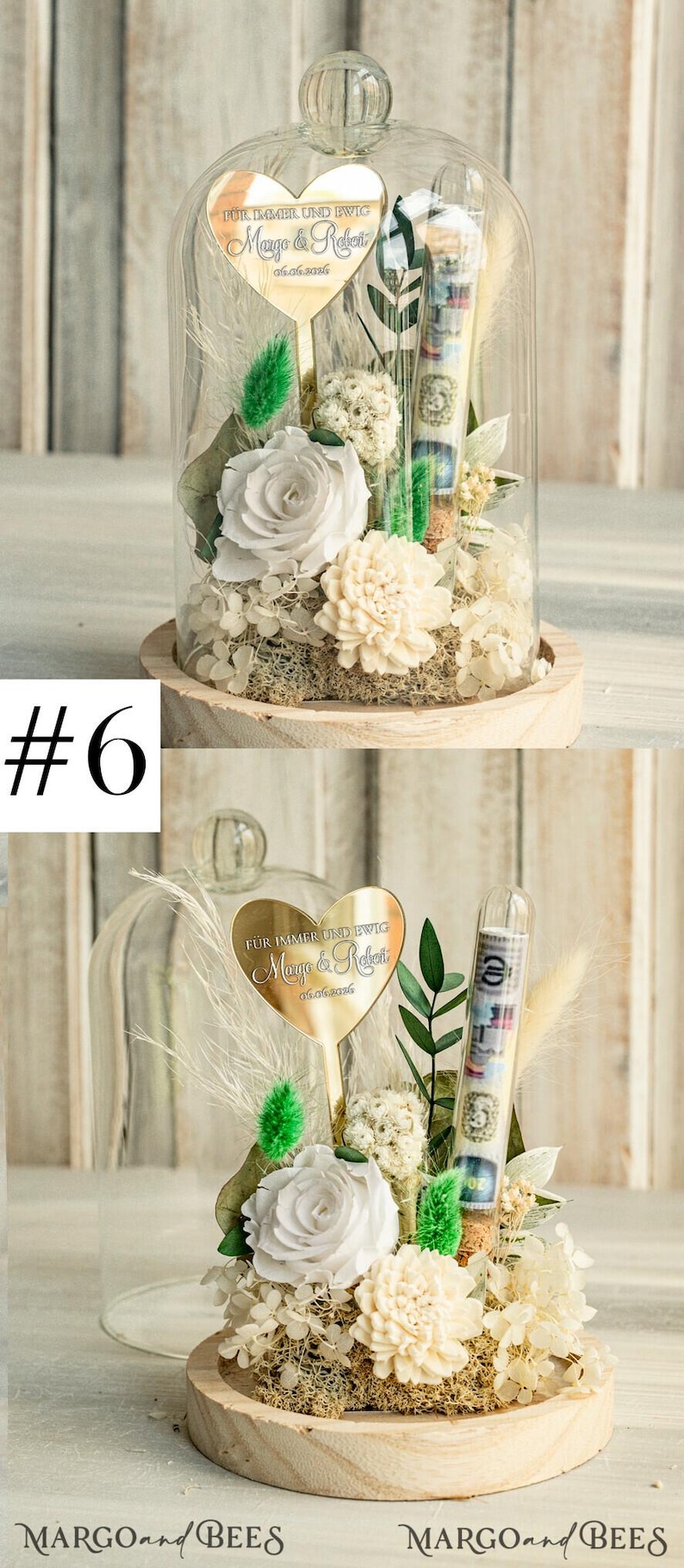 May include: A glass dome with a wooden base contains a white rose, white carnations, and green foliage. A gold heart-shaped plaque with the text "F&Uuml;R IMMER UND EWIG Mango & Relet 06.06.2016" is attached to a stick in the center of the dome. A clear glass tube filled with money is also inside the dome.