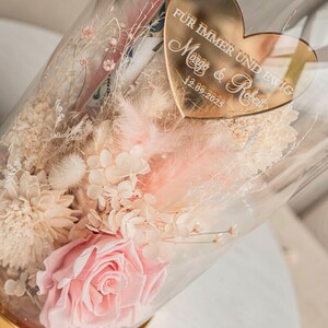 May include: A glass dome filled with dried flowers, including a pink rose, white hydrangeas, and white pampas grass. A gold heart-shaped plaque with the text "F&uuml;r immer und ewig Margo & Robert 12.09.2023" is attached to the dome.