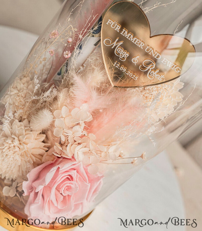 May include: A glass dome filled with dried flowers, including a pink rose, white hydrangeas, and white pampas grass. A gold heart-shaped plaque with the text "F&uuml;r immer und ewig Margo & Robert 12.09.2023" is attached to the dome.