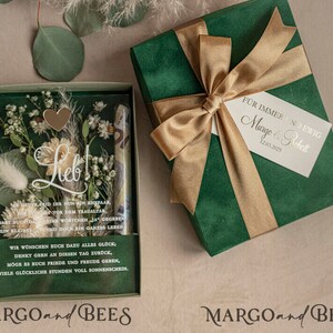 May include: A green gift box with a gold ribbon and a white tag that says "For always and forever, Margo & Robert 12.03.2023". The box is open and inside is a clear box with dried flowers and a heart-shaped charm. The text "Lieb!" is written on the box. The box is labeled "MARGO and BEES".