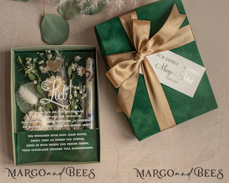 May include: A green gift box with a gold ribbon and a white tag that says "For always and forever, Margo & Robert 12.03.2023". The box is open and inside is a clear box with dried flowers and a heart-shaped charm. The text "Lieb!" is written on the box. The box is labeled "MARGO and BEES".