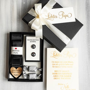 May include: A black gift box with a white satin ribbon and bow. The box contains a black tie, cufflinks, and two small candies. The box is labeled "Liebster Papa" and includes a card with the text "Liebster Papa Du warst der erste Mann, den ich geliebt habe und f&uuml;r immer lieben werde. HEUTE BRAUT, MORGEN EHEFRAU ABER F&Uuml;R IMMER DEINE TOCHTER Deine Nelle 25.05.2025"