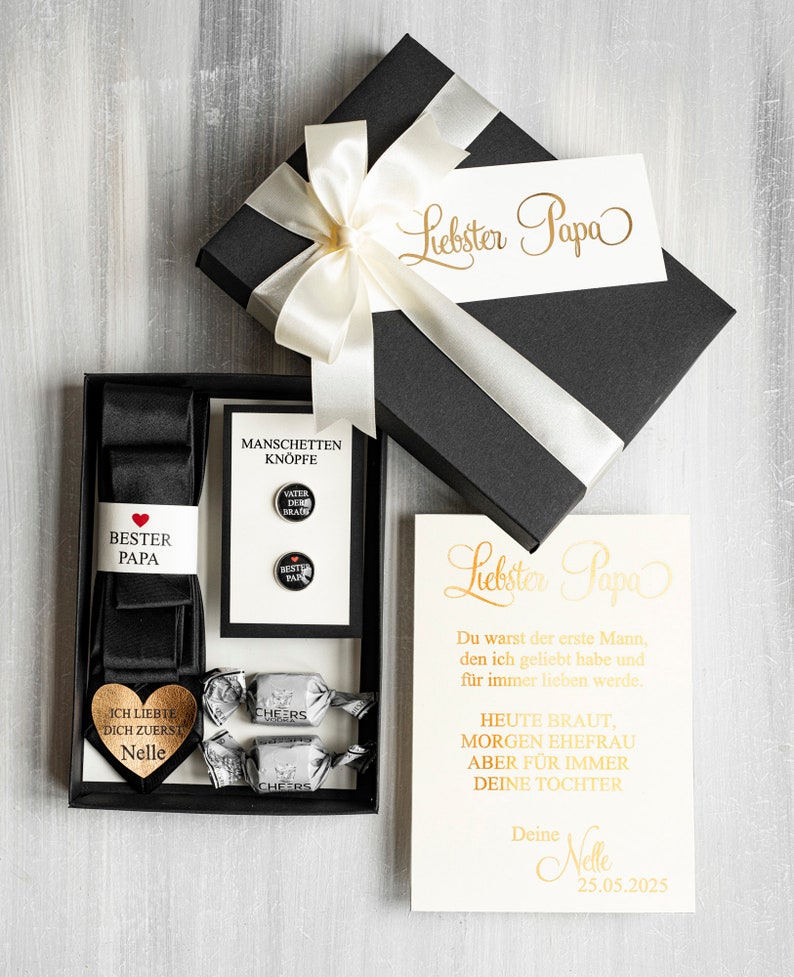 May include: A black gift box with a white satin ribbon and bow. The box contains a black tie, cufflinks, and two small candies. The box is labeled "Liebster Papa" and includes a card with the text "Liebster Papa Du warst der erste Mann, den ich geliebt habe und f&uuml;r immer lieben werde. HEUTE BRAUT, MORGEN EHEFRAU ABER F&Uuml;R IMMER DEINE TOCHTER Deine Nelle 25.05.2025"