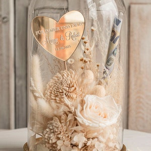 Op de afbeelding: Een glazen stolp met een gouden voet, die een boeket gedroogde bloemen bevat, waaronder een witte roos, wit pampasgras en witte chrysanten. Een gouden hartvormige plaquette met de tekst "F&uuml;r immer und ewig" en de namen "Marco & Rosa" en de datum "12.09.2023" is in het midden van het boeket geplaatst.