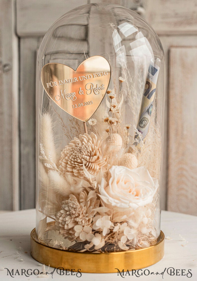 Op de afbeelding: Een glazen stolp met een gouden voet, die een boeket gedroogde bloemen bevat, waaronder een witte roos, wit pampasgras en witte chrysanten. Een gouden hartvormige plaquette met de tekst "F&uuml;r immer und ewig" en de namen "Marco & Rosa" en de datum "12.09.2023" is in het midden van het boeket geplaatst.