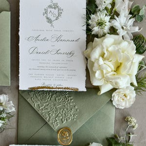May include: A rectangular wedding invitation with deckled edges and gold foil accents at the top and bottom. The text features a floral crest with the initials A/D, followed by names and event details. It is paired with a sage green envelope featuring an embossed floral pattern and a gold wax seal.