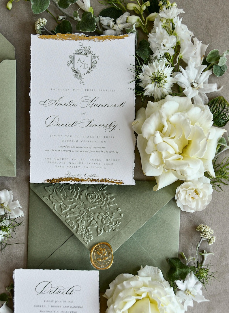 May include: A rectangular wedding invitation with deckled edges and gold foil accents at the top and bottom. The text features a floral crest with the initials A/D, followed by names and event details. It is paired with a sage green envelope featuring an embossed floral pattern and a gold wax seal.