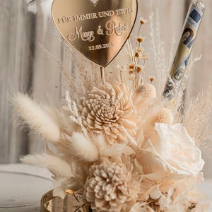 Op de afbeelding: Een gouden hartvormige plaquette met de tekst "F&uuml;r immer und ewig Margo & Robert 12.09.2023" zit bovenop een glazen stolp gevuld met gedroogde bloemen en een glazen buis met geld.