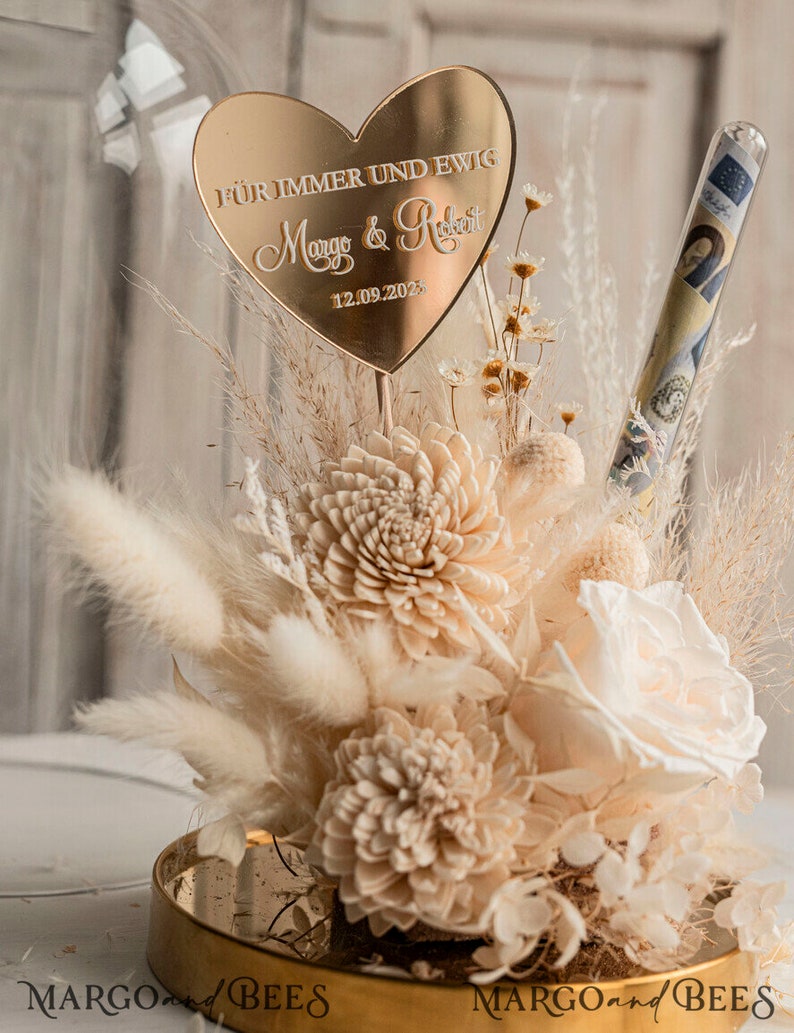 Op de afbeelding: Een gouden hartvormige plaquette met de tekst "F&uuml;r immer und ewig Margo & Robert 12.09.2023" zit bovenop een glazen stolp gevuld met gedroogde bloemen en een glazen buis met geld.