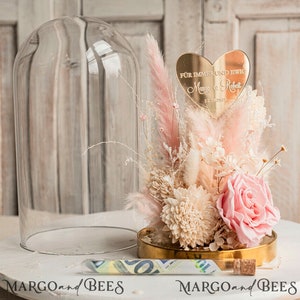 May include: A glass dome cloche with a pink and white dried flower arrangement. The arrangement includes a pink rose, white dried flowers, and pink pampas grass. A gold heart-shaped plaque with the text "F&uuml;r immer und ewig" and the names "Maya & Robert" and the date "12.09.2025" is placed on top of the arrangement.