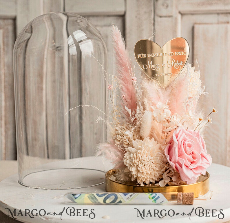 May include: A glass dome cloche with a pink and white dried flower arrangement. The arrangement includes a pink rose, white dried flowers, and pink pampas grass. A gold heart-shaped plaque with the text "F&uuml;r immer und ewig" and the names "Maya & Robert" and the date "12.09.2025" is placed on top of the arrangement.