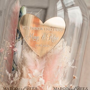 May include: A gold heart-shaped plaque with the text "F&uuml;r immer und ewig Margo & Robert 12.09.2025" is displayed inside a glass dome with dried flowers.