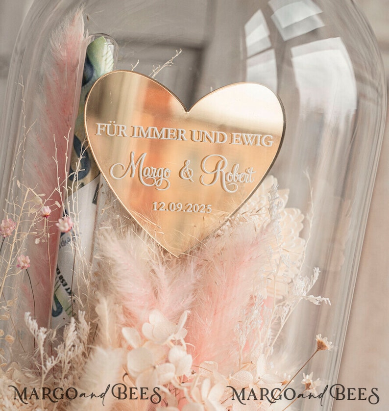 May include: A gold heart-shaped plaque with the text "F&uuml;r immer und ewig Margo & Robert 12.09.2025" is displayed inside a glass dome with dried flowers.